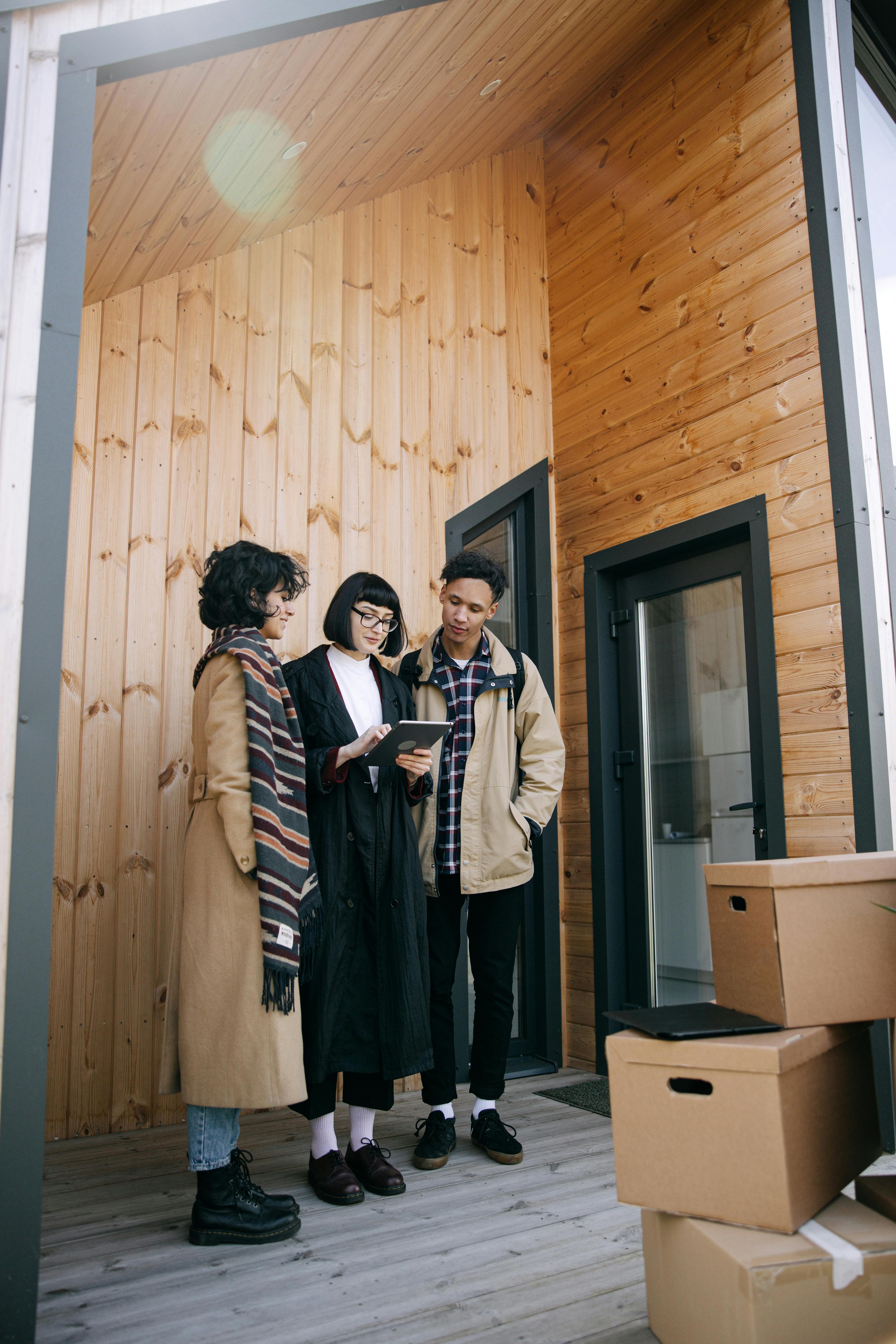 Three young adults standing with moving boxes outside a modern wooden house, using a tablet.