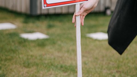 Close-up of a red 'House for Rent' sign on a grassy lawn, hand holding pole.