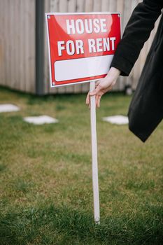 Close-up of a red 'House for Rent' sign on a grassy lawn, hand holding pole.