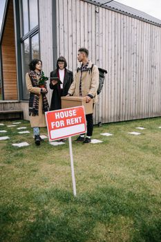 Three adults viewing a wooden house for rent with a sign on grassy yard.