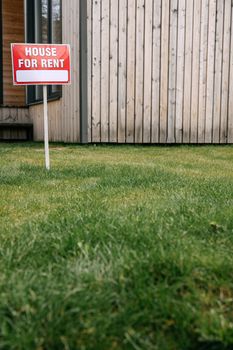 Red 'House for Rent' sign on green lawn with wooden house wall in background.
