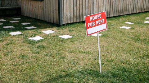 A red 'House for Rent' sign stands on a grassy lawn beside a wooden house exterior.