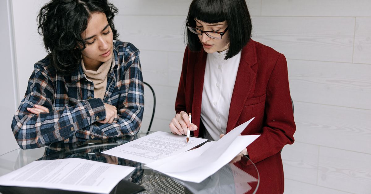 Two women collaborating at a glass table, reviewing business documents in a modern office setting.