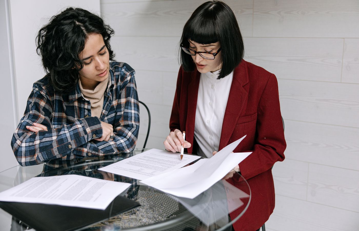 Two women collaborating at a glass table, reviewing business documents in a modern office setting.