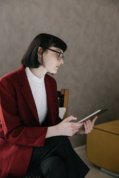 Young woman wearing red blazer and eyeglasses using a tablet while sitting indoors.