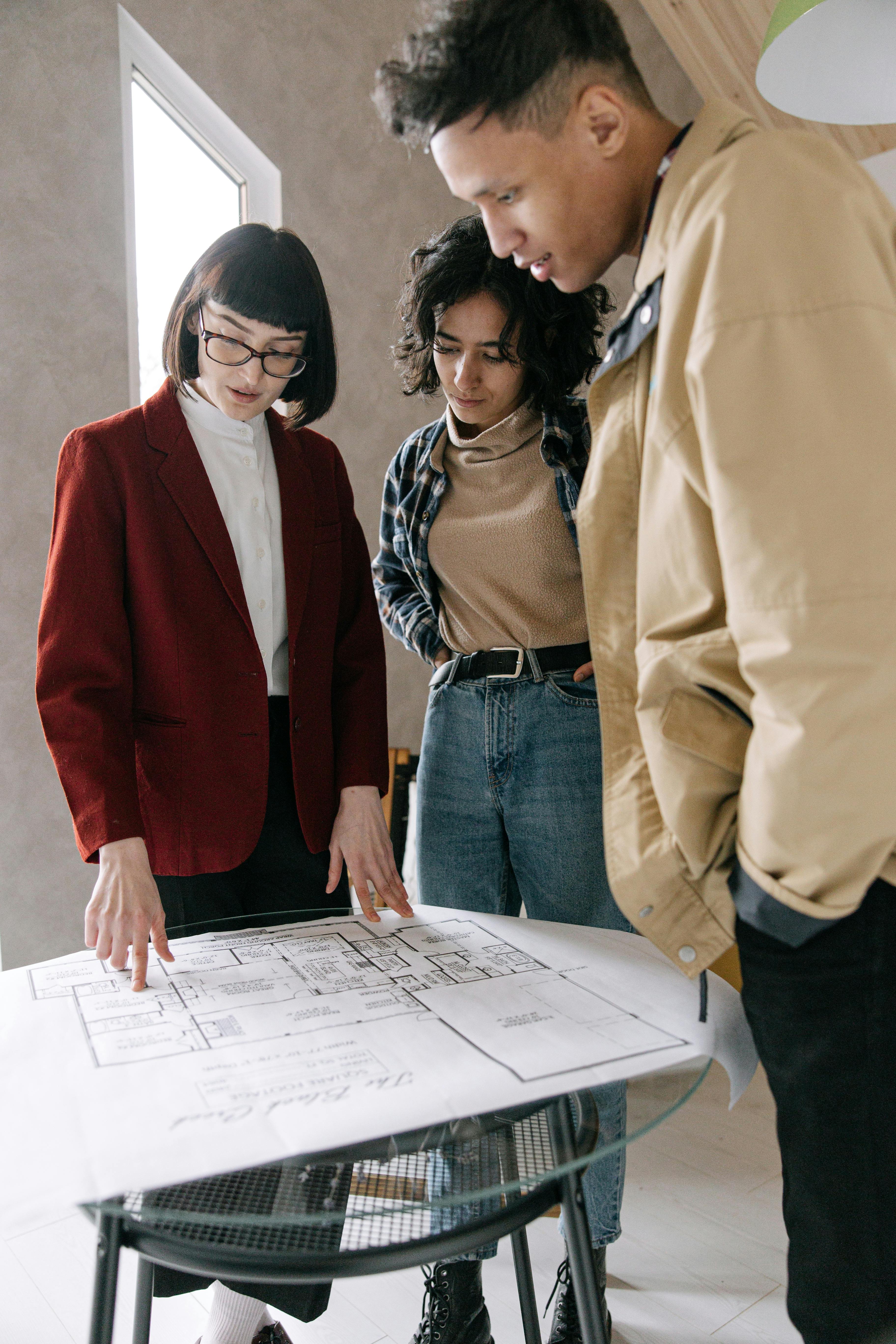 couple standing beside woman in red blazer pointing at blueprint