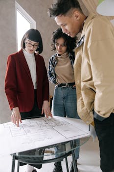 A real estate agent discussing blueprints with a young couple at a glass table indoors.