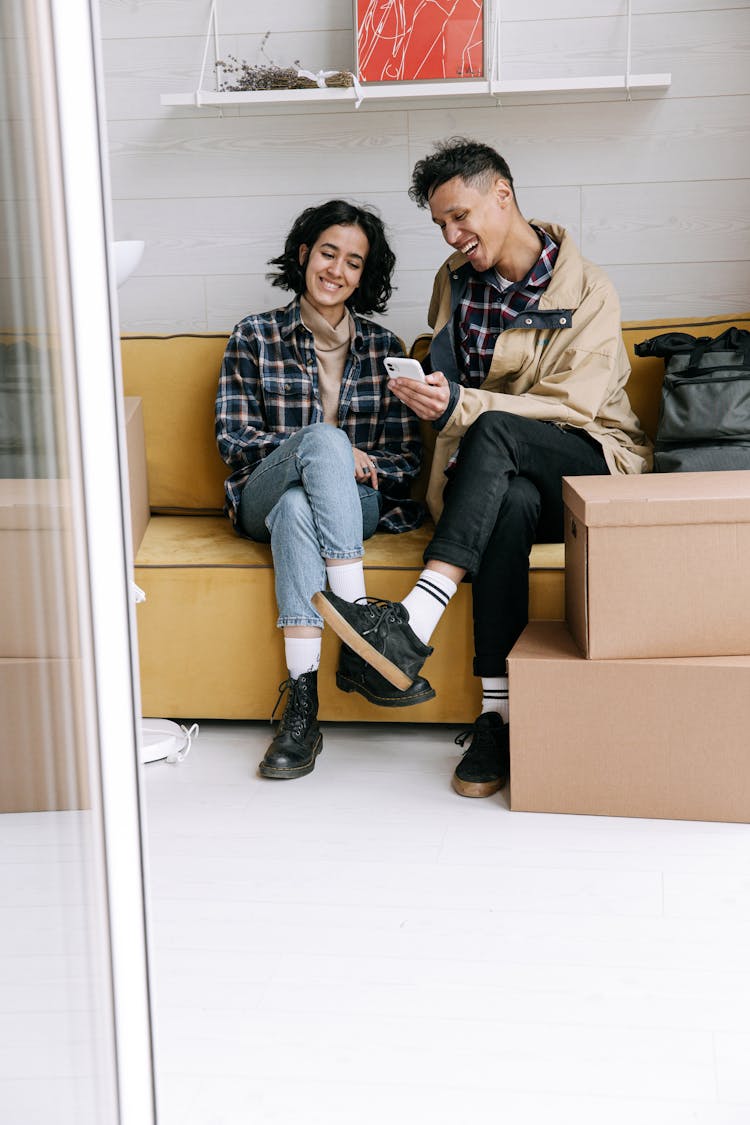 Young Couple Sitting On A Sofa In Their New Home And Looking At A Smartphone Screen 