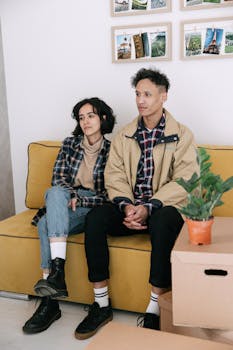 Casually dressed young couple sitting on a yellow sofa with moving boxes nearby.