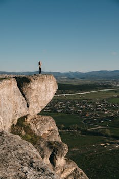 A woman stands on a cliff edge with a panoramic view of the rural landscape below, under clear blue skies.