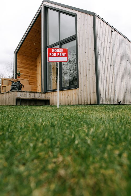 Free A modern wooden house with a 'for rent' sign on a lush green lawn. Stock Photo