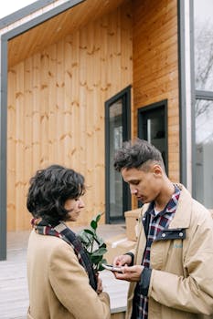 Young couple standing outside their new wooden home holding a plant and smartphone.