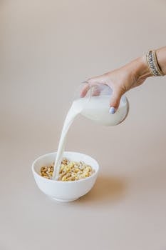 Close-up of a hand pouring milk into a bowl of cereal, showcasing breakfast preparation.