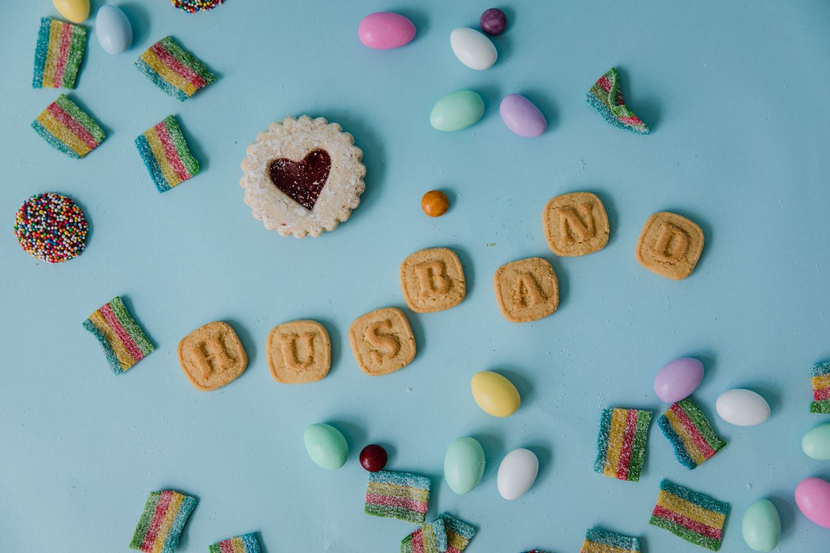 Playful candy flat lay with cookies spelling 'Husband' on a pastel blue backdrop.