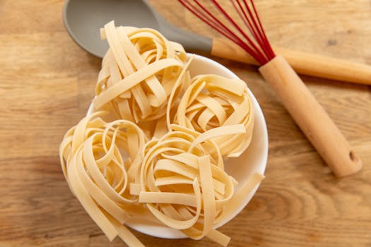 High-angle view of uncooked tagliatelle pasta in a bowl with kitchen utensils on a wooden surface.