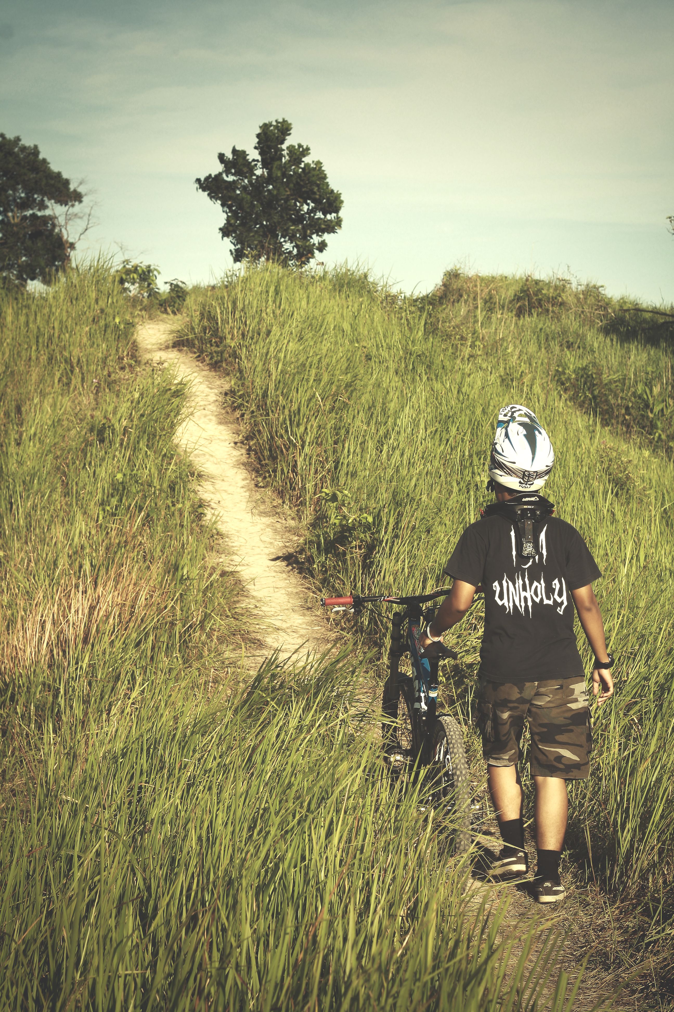 A mountain biker with a helmet walks on a grassy path under clear skies.