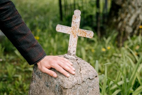 Free A detailed view of a hand resting on a rusty metal cross in a serene, grassy cemetery. Stock Photo