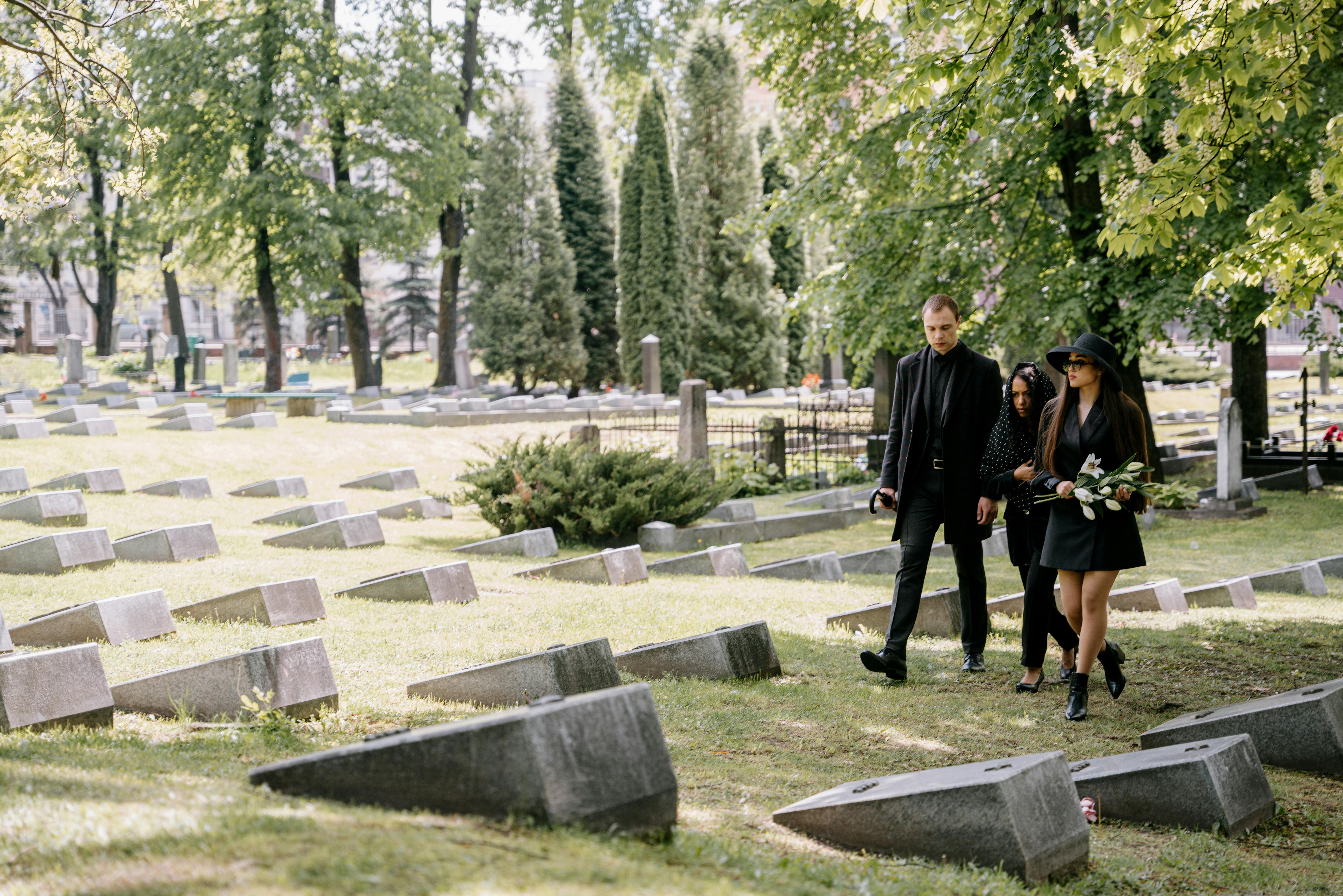 Mourners walk through a serene cemetery surrounded by lush green trees, showing respect and reflection.