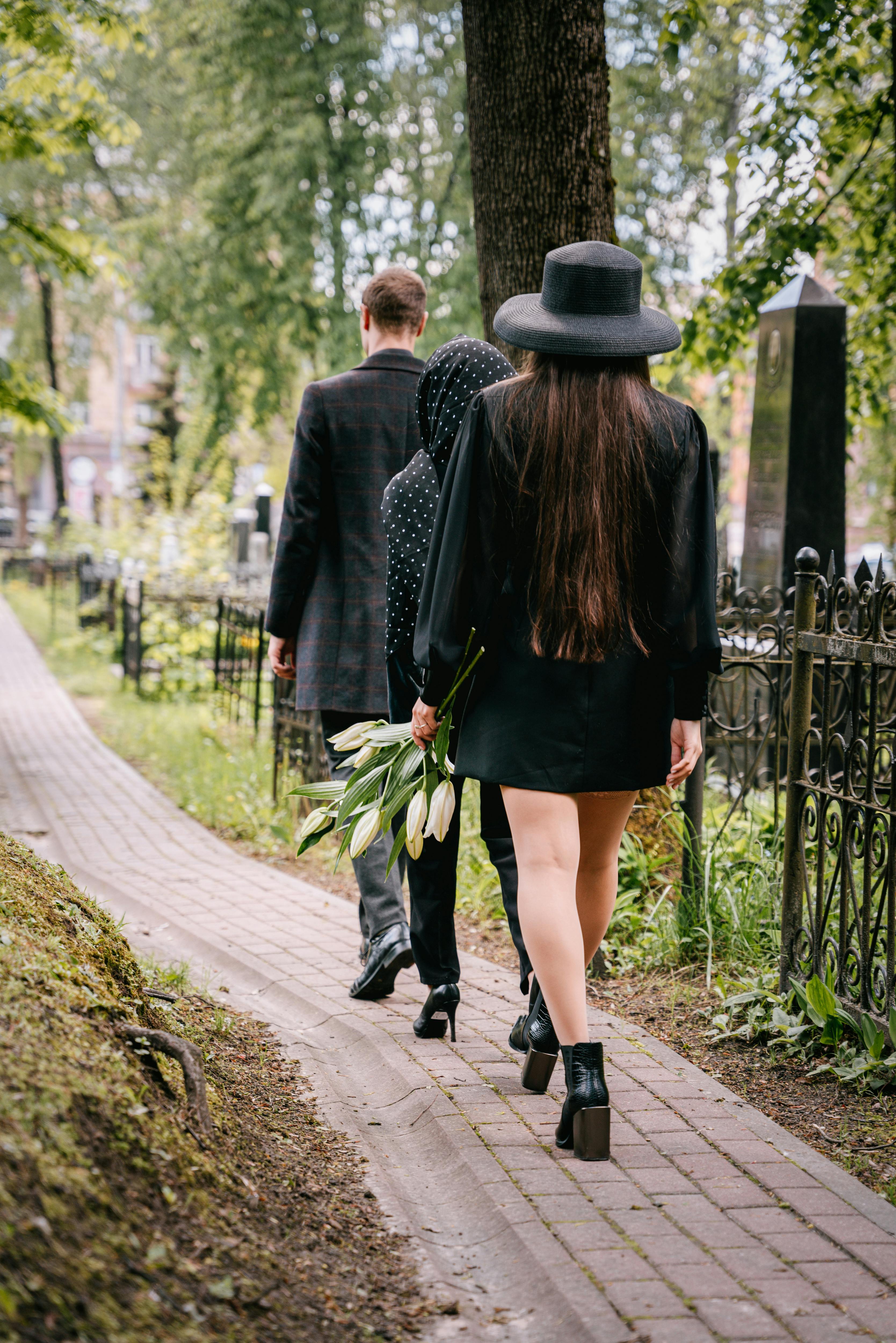 Elegant mourners in black walk through a serene cemetery holding flowers, symbolizing loss and remembrance.