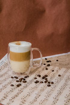 A glass of layered latte on sheet music with scattered coffee beans, perfect for a cozy ambiance.