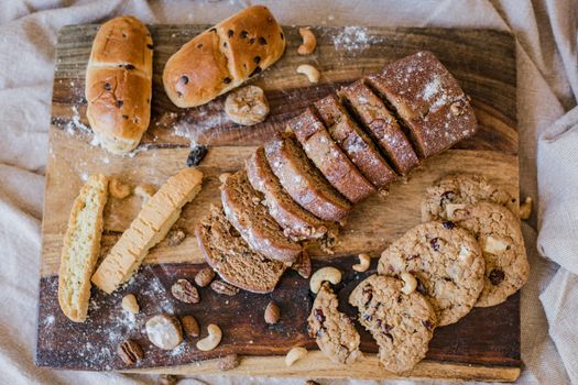 Overhead view of sliced bread, cookies, and pastries on a wooden board, styled with nuts.