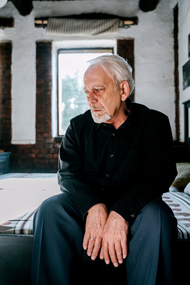 An Elderly Man In Black Long Sleeves Sitting On The Bed With A Sad Face