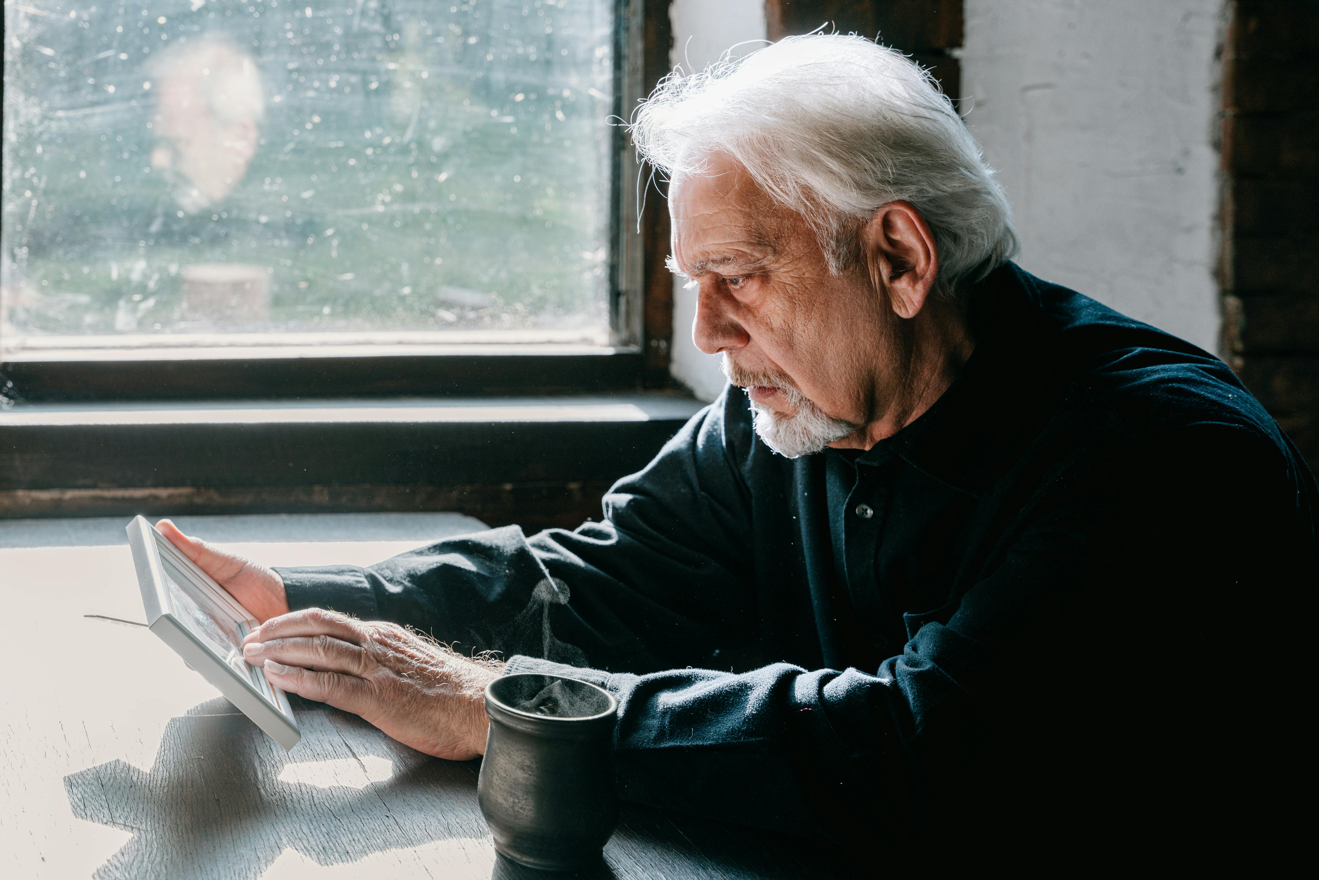 Senior man with gray hair sits by the window, holding a photo, conveying deep reflection.
