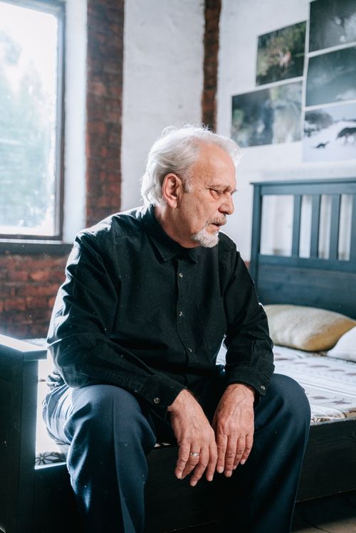 Free An elderly man in a black shirt sits pensively on a bed in a bright bedroom. Stock Photo