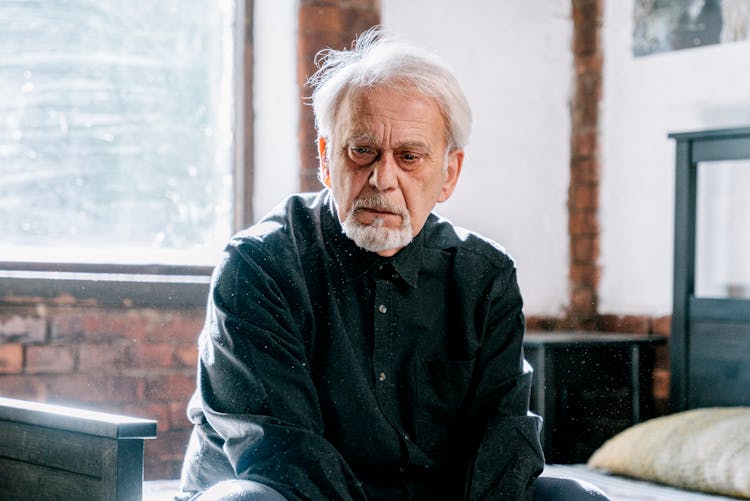 An Elderly Man In Black Long Sleeves Sitting On The Bed