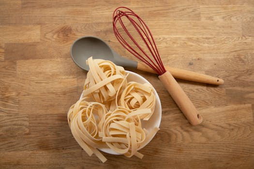 Flat lay of uncooked tagliatelle pasta with a whisk and spoon on a wooden surface.