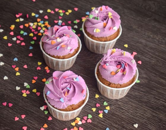 Four vanilla cupcakes with swirled purple buttercream and heart sprinkles on dark wood