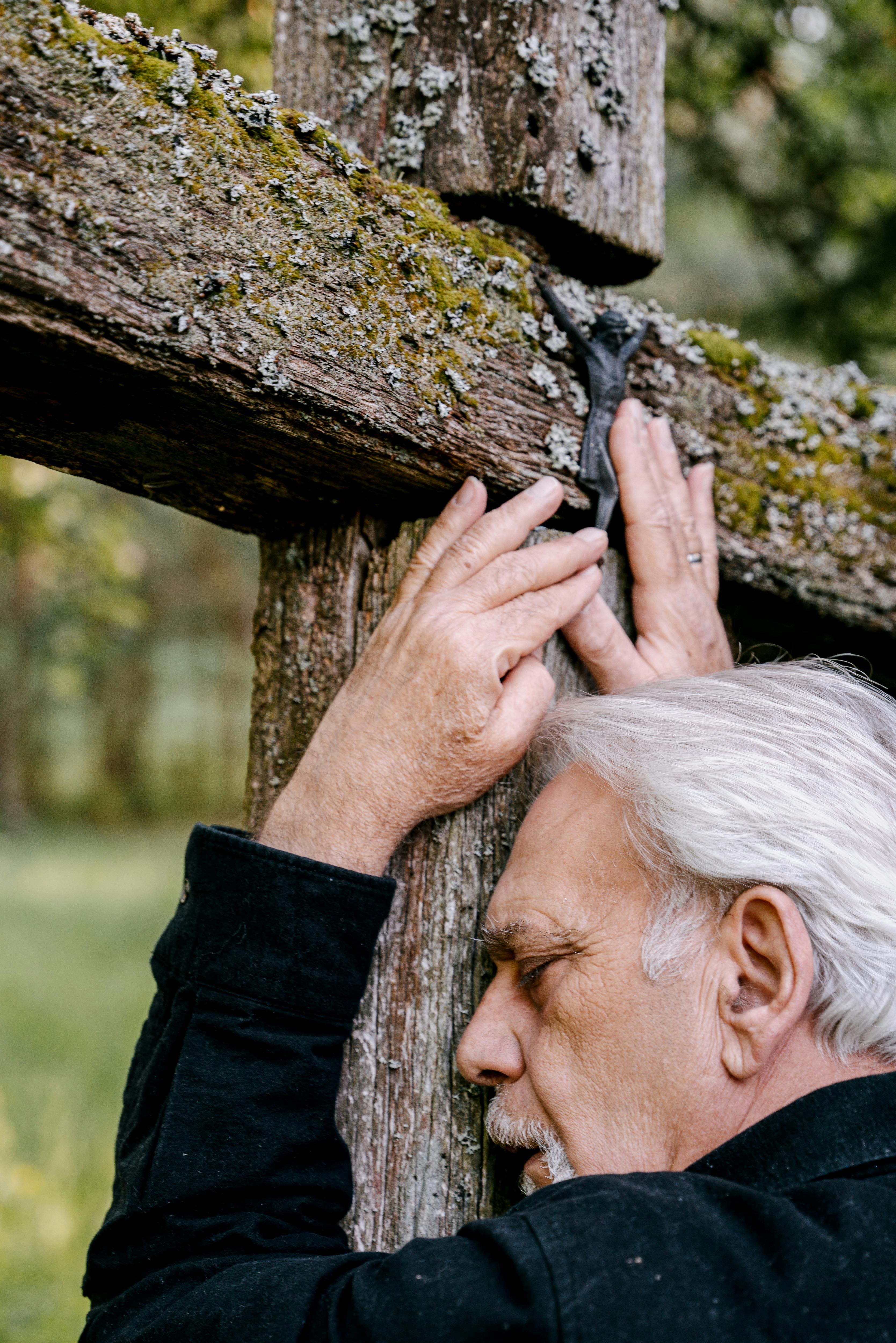 Elderly man mourns at a moss-covered wooden cross in a serene graveyard.
