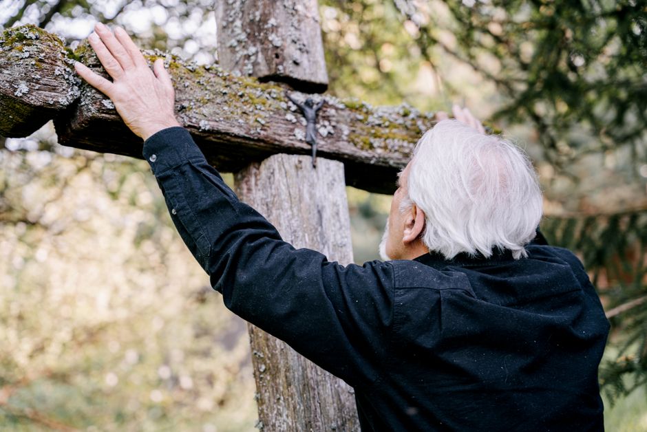 Senior man in black shirt holding a wooden cross in a forest setting, symbolizing grief and reflection.