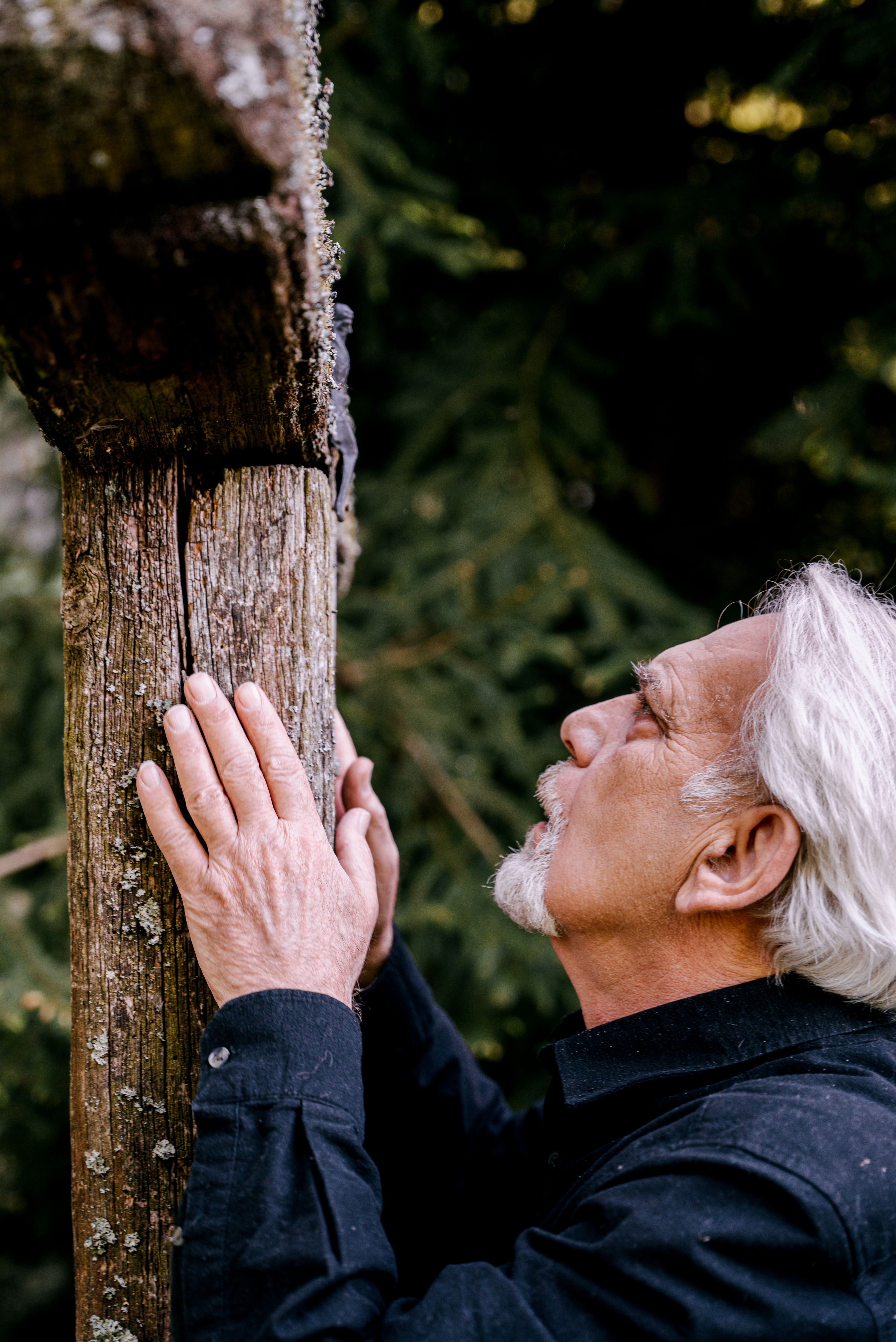 An elderly man with white hair reflects at a wooden cross in an outdoor setting, conveying a sense of loss and reverence.