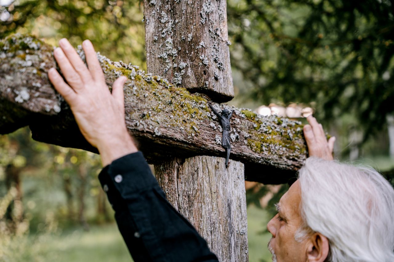 an elderly man reflects by a moss covered wooden cross in a serene out