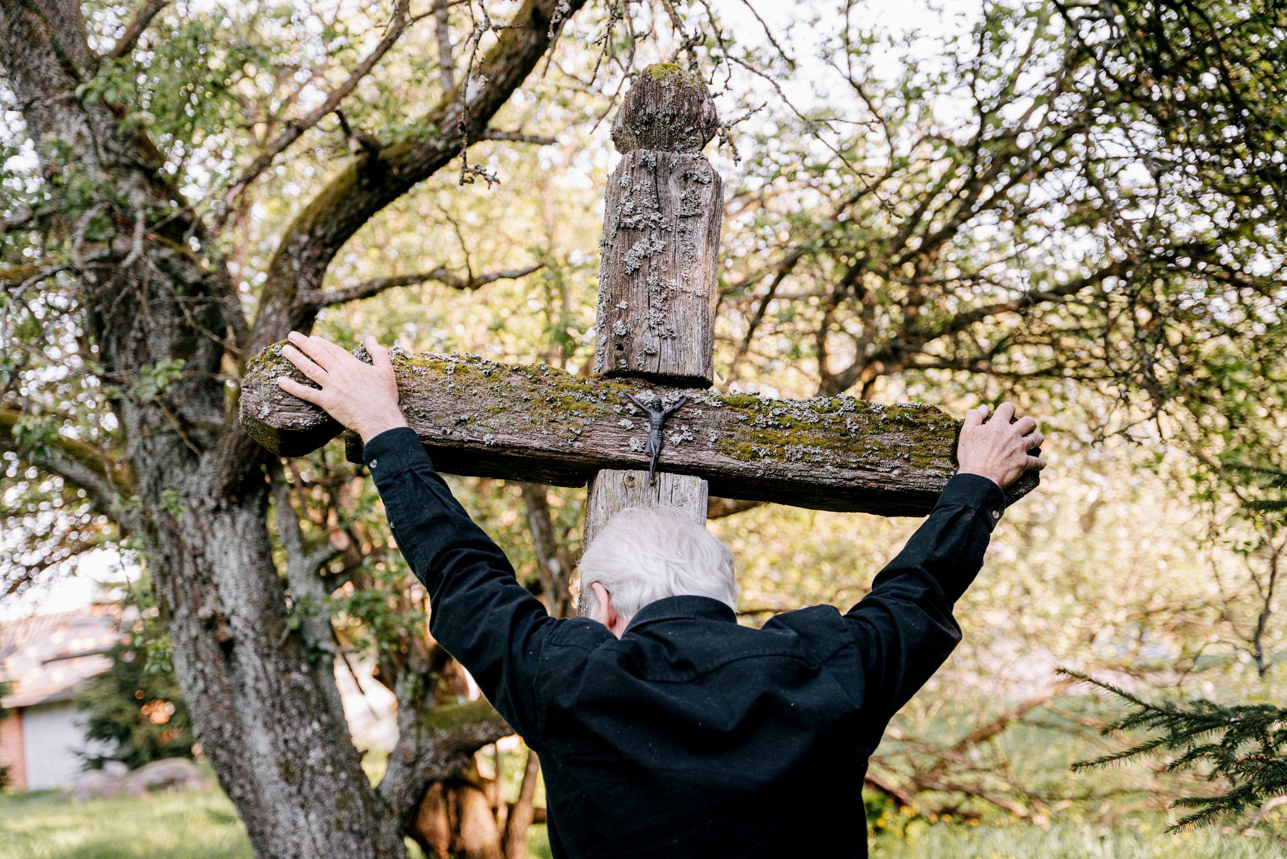 An elderly man embraces a wooden cross, expressing grief and religion in an outdoor setting.