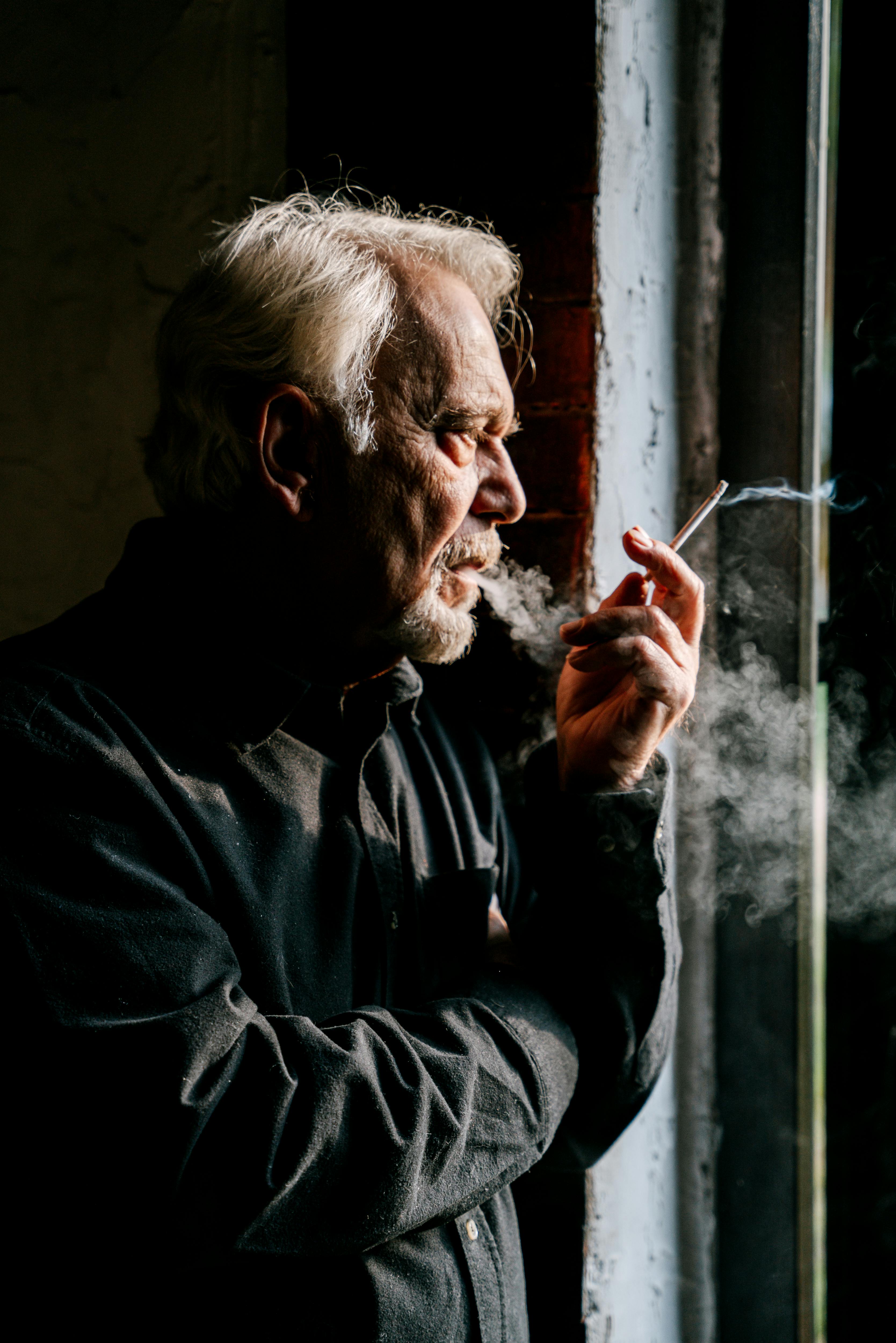 A senior man with white hair smokes thoughtfully by a window, casting dramatic shadows.
