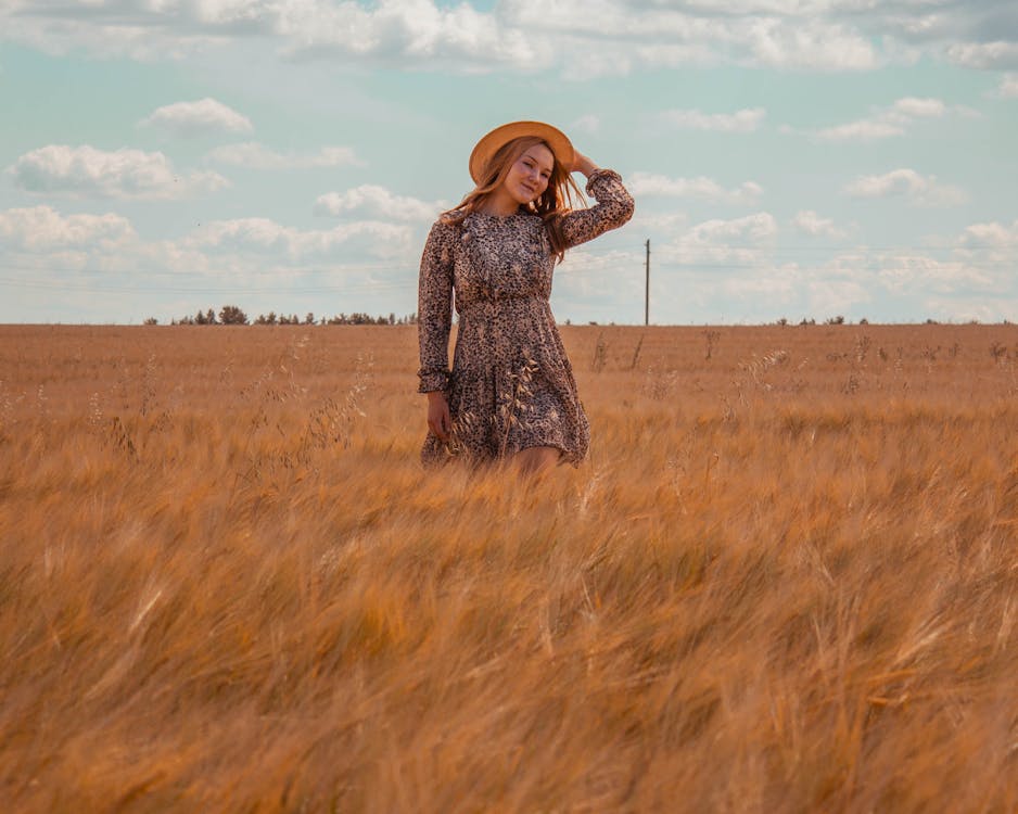 Free A Woman in Floral Dress Wearing a Hat while Standing on the Grass Field Stock Photo