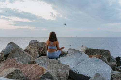 Free A woman meditates on rocky beach during sunrise, promoting relaxation and mindfulness. Stock Photo