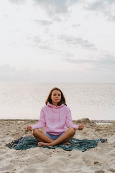 A woman in a pink hoodie meditates on a beach, embodying relaxation and mindfulness by the sea.
