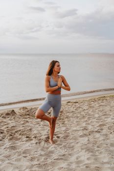 A woman performs yoga's tree pose on a sandy beach during daylight, embracing nature and calmness.