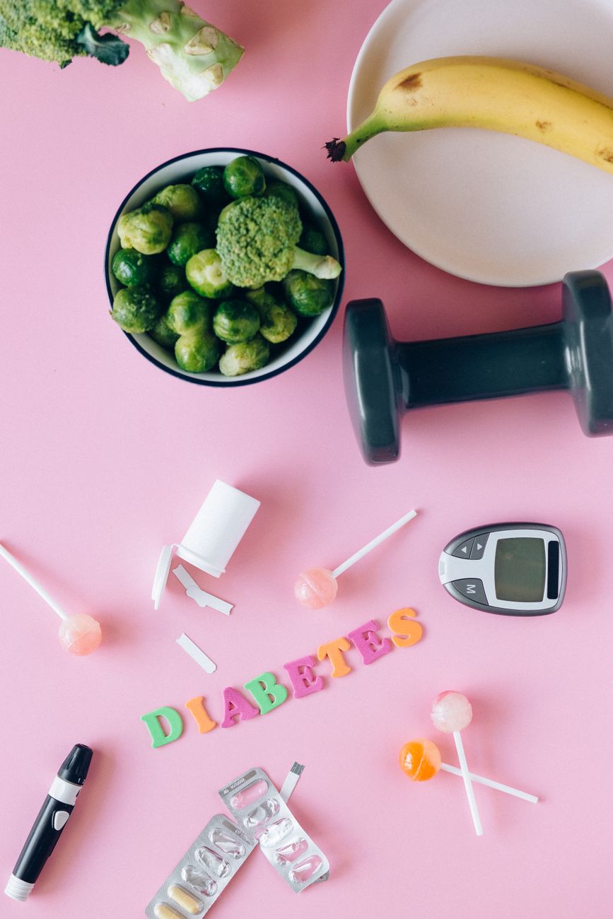 Flat lay of diabetes and health management items including fruits, glucometer and a dumbbell on a pink background.