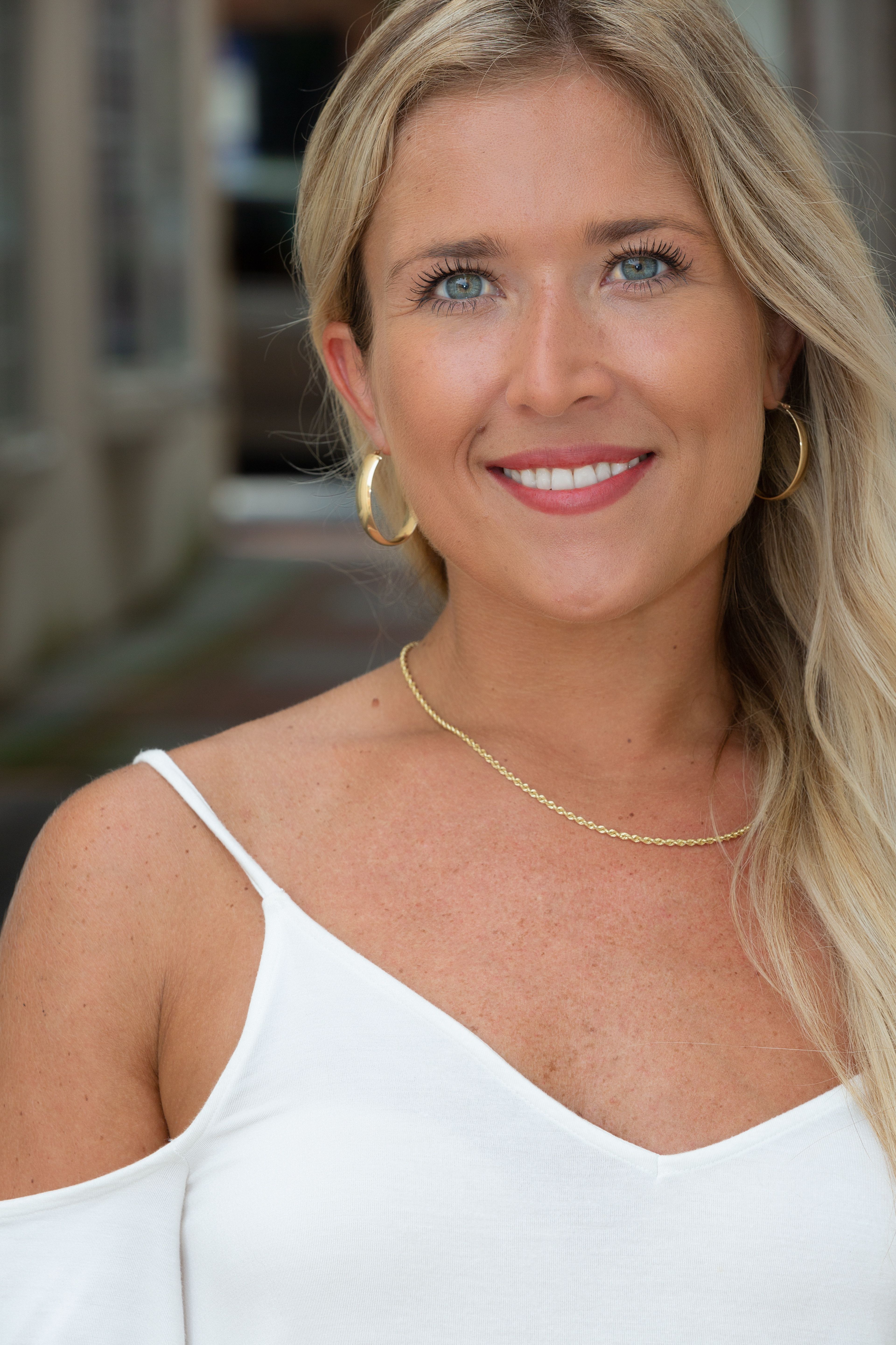 Portrait of a smiling woman wearing a white top and gold jewelry outdoors.