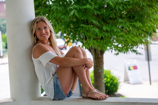 Happy blonde woman wearing a white top and denim shorts, sitting outdoors on a sunny day.