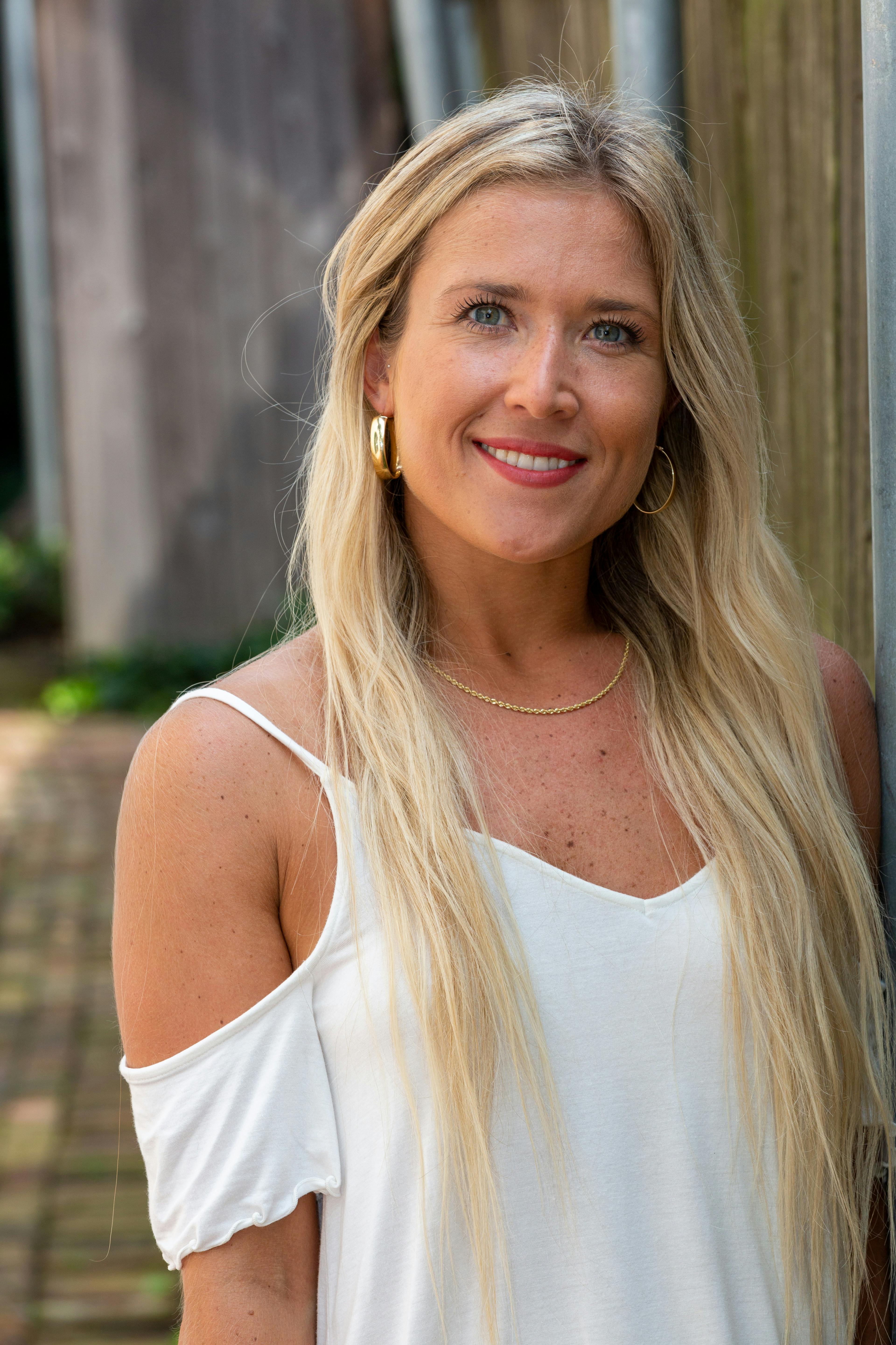 Portrait of a smiling blonde woman in a white top with loop earrings outdoors.