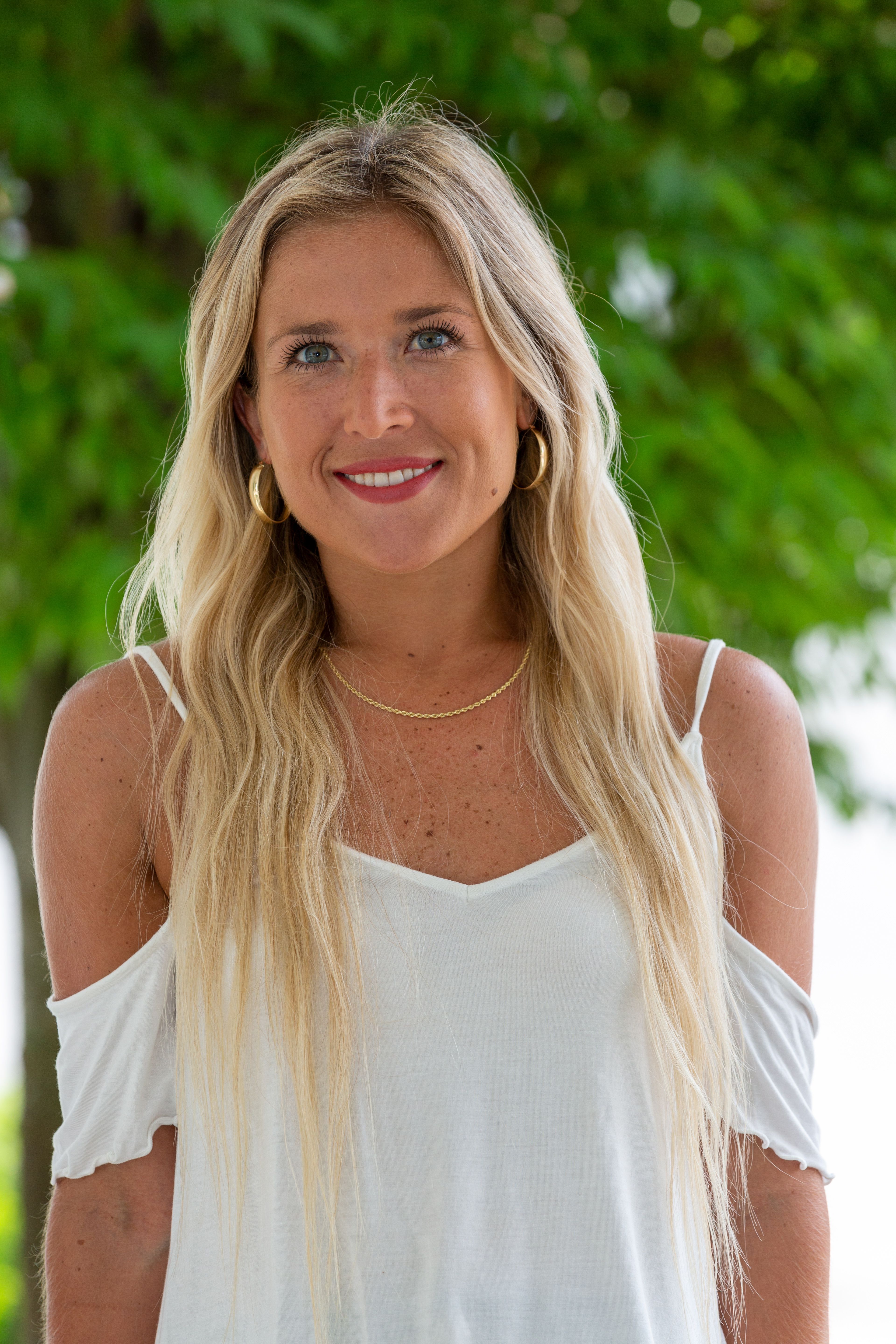 Portrait of a smiling blonde woman with long hair and white top outdoors.
