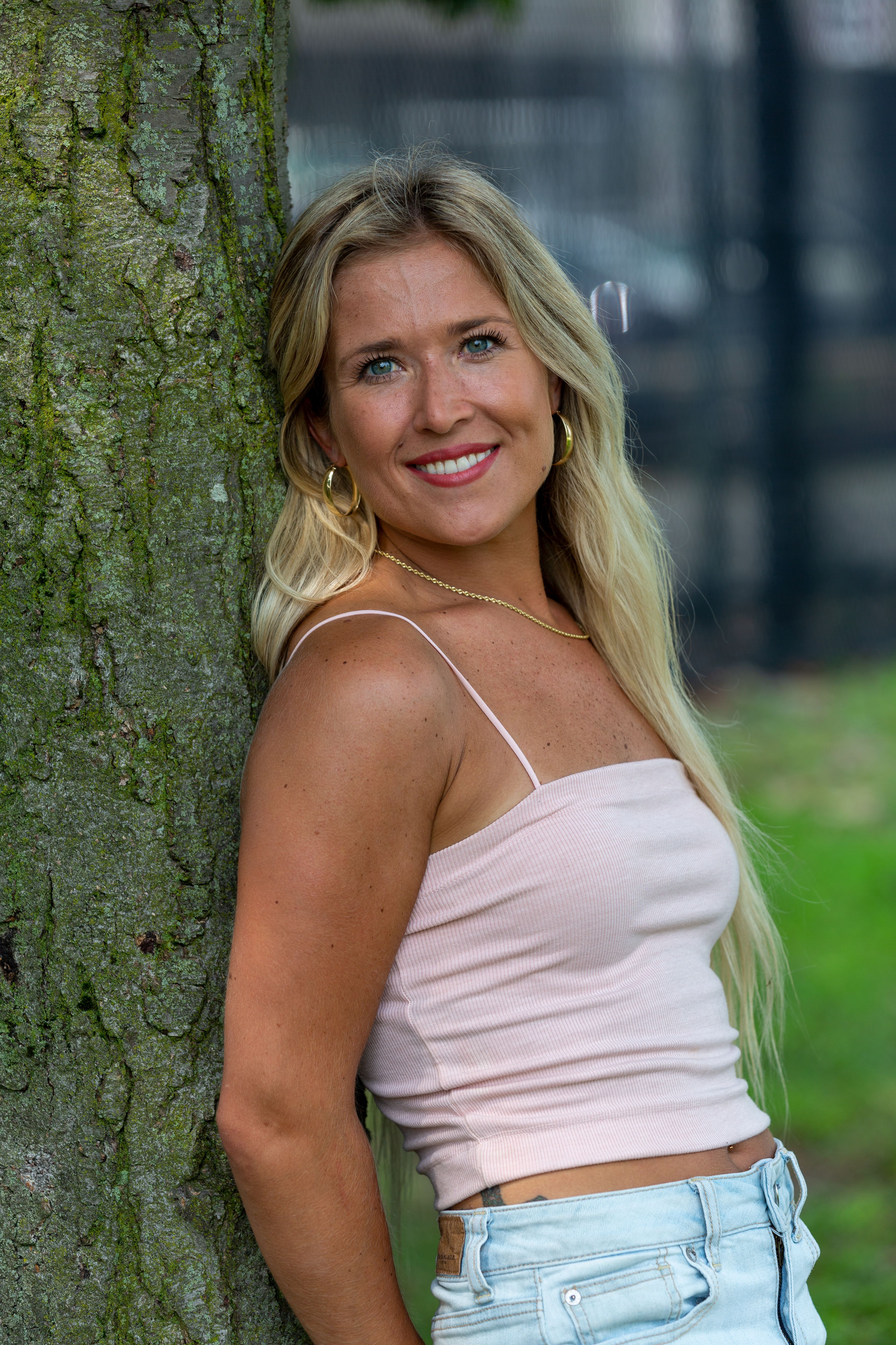 Blonde woman in light top and shorts, smiling while leaning against a tree.