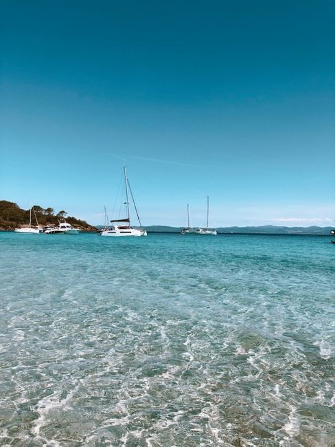 A tranquil beach view featuring sailboats docked on calm, clear waters under a bright blue sky.