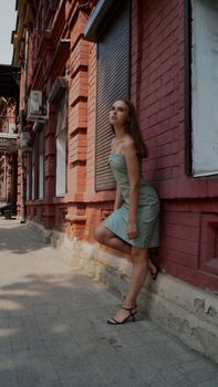 Woman in green dress posing against a brick wall with foot raised, creating an elegant side view.