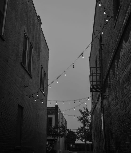 Black and white photo of an alleyway with hanging string lights, evoking a moody urban atmosphere.